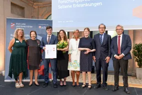 Acht Personen stehen festlich gekleidet auf einer Bühne - Frauen und Männer - die 3. Person von links hält eine Urkunde in der Hand. Im Hintergrund Banner und Logo vom Eva Luise köhler Forschungspreis 2025.