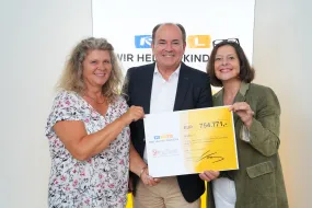 2 lachende Frauen haben einen läterern Herrn auch lachend in ihre MItte genommen und halten gemeinsam einen Scheck hoch. Auf dem Scheck steht die Summe 754771 Euro, das Logo von RTL Wir helfen Kindern und der ACHSE ist ebenfalls drauf. 