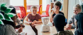 Kinder sitzen an einem Tisch, in der Mitte steht ein kleiner Roboter. 