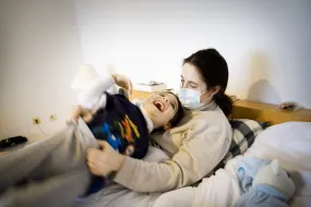 Mutter mit Mund-Nasen-Schutz liegt im Bett und hält lachendes Kind auf dem Arm. Das Kind hat einen seltenen Gendefekt. 