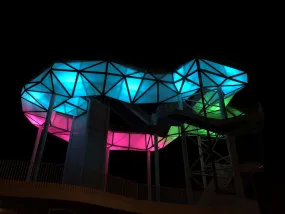 Auf dem Bild ist ein architektonisch außergewöhnliches Gebäude aus Glas und Stahl zu erkennen, das in den Farben blau, pink und grün angestrahlt ist.