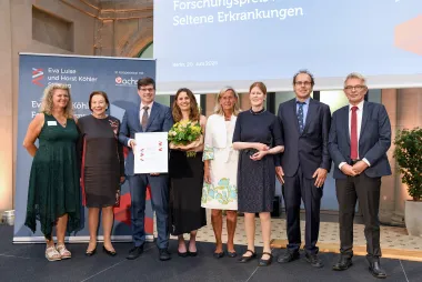 Acht Personen stehen festlich gekleidet auf einer Bühne - Frauen und Männer - die 3. Person von links hält eine Urkunde in der Hand. Im Hintergrund Banner und Logo vom Eva Luise köhler Forschungspreis 2025.