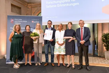 7 Menschen auf einer Bühne, festlich gekleidet, Männer und Frauen, die vierte Person von link hält eine Urkunde in die Kamera und freut sich. Im Hintergrund Banner und Logo vom Forschungspreis 2025.