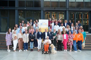 Viele Menschen stehen bunt gekleidet und lächelnd vor einem gläsernen Gebäude. Ein Banner mit der Aufschrift RTL-Wir helfen Kindern. 