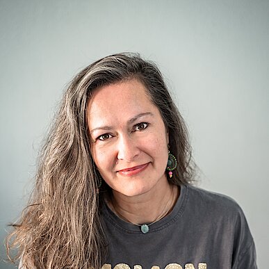 Kati Birr Frau mit langen, gewellten Haaren trägt ein dunkelgraues T-Shirtlgraues T-Shirt mit goldener Aufschrift 'MAMAN D'AMOUR'