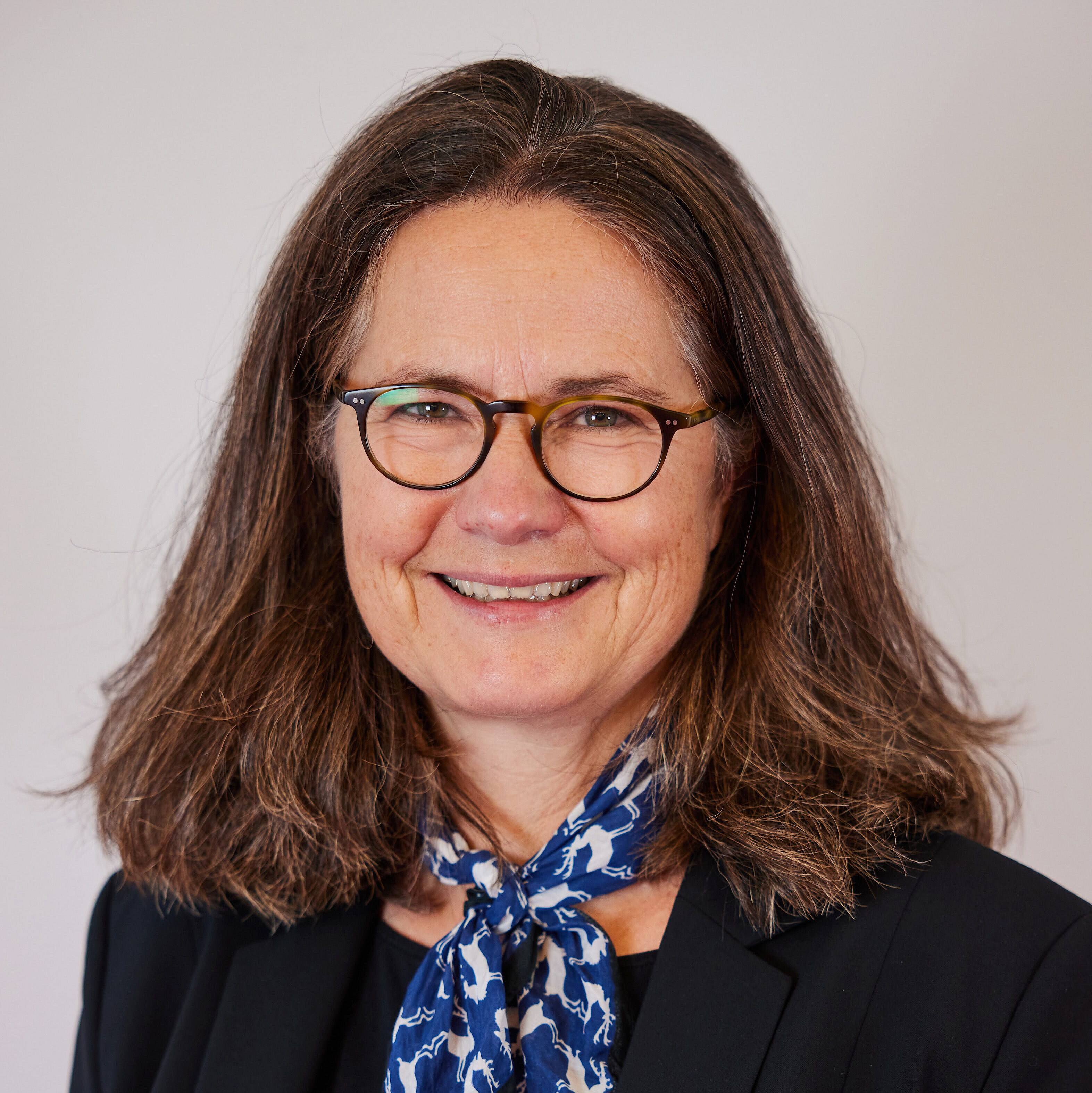 Frau mit schulterlangem braunem Haar, schwarzem Blazer, schwarzem Oberteil und blau-weiß gemustertem Halstuch.