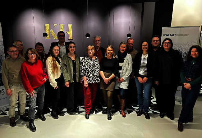 Gruppenfoto von Menschen in einem Raum mit dunkler Wand und goldenen Buchstaben KH im Hintergrund