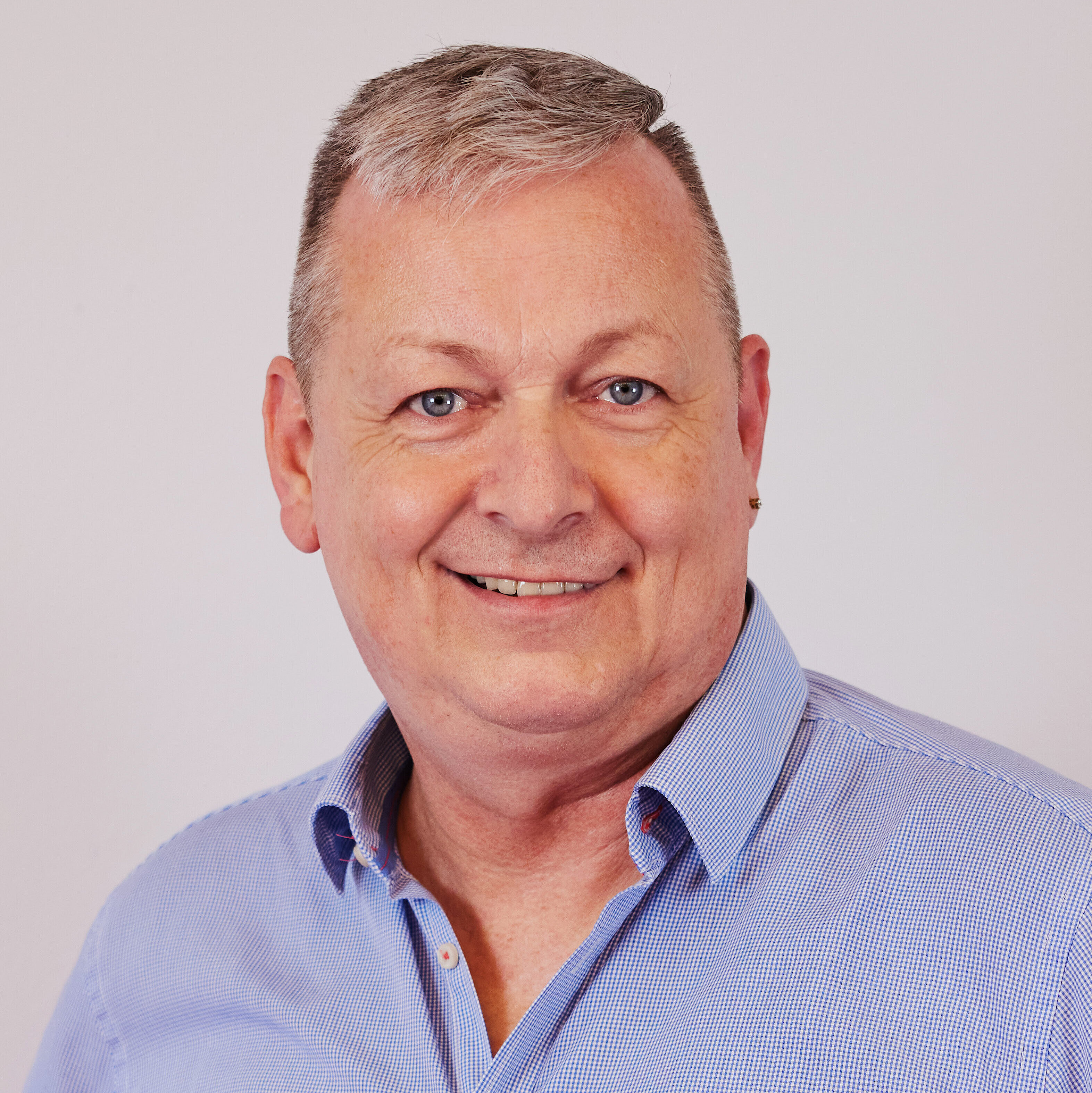 Mann mit grauen Haaren trägt hellblaues Hemd mit offenem Kragen vor einfarbigem Hintergrund.