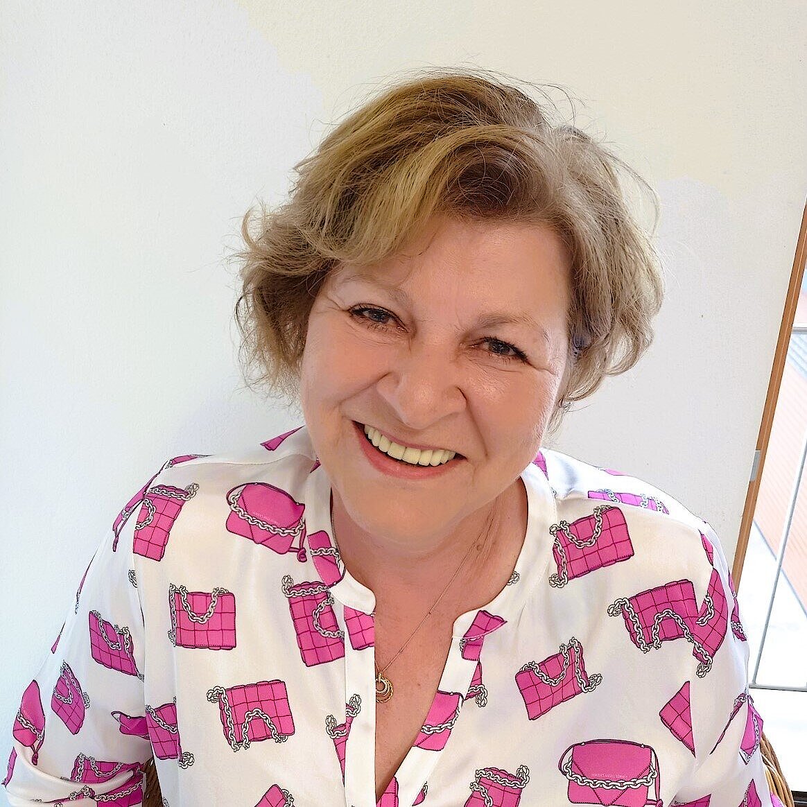 Eine Frau mit blondem, halblangem Haar sitzt auf einem Stuhl vor einer weißen Wand. Sie trägt eine weiße Bluse mit auffälligem Muster aus pinkfarbenen Handtaschen mit Kettendetails. Im Hintergrund ist ein Fenster mit hellem Tageslicht zu sehen.