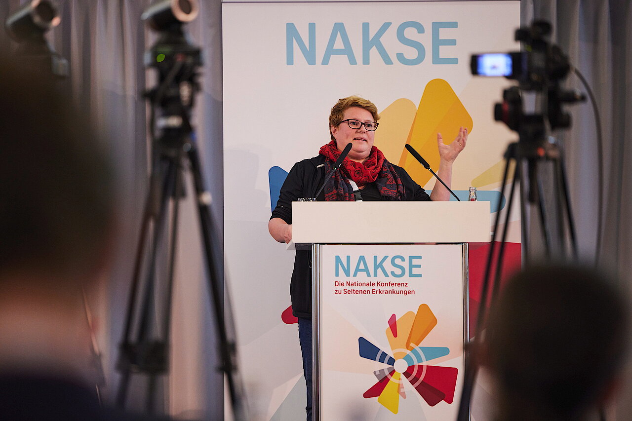 Person mit rotem Halstuch steht an einem Rednerpult mit Mikrofon vor einem Banner mit dem Schriftzug 'NAKSE' und buntem Logo.