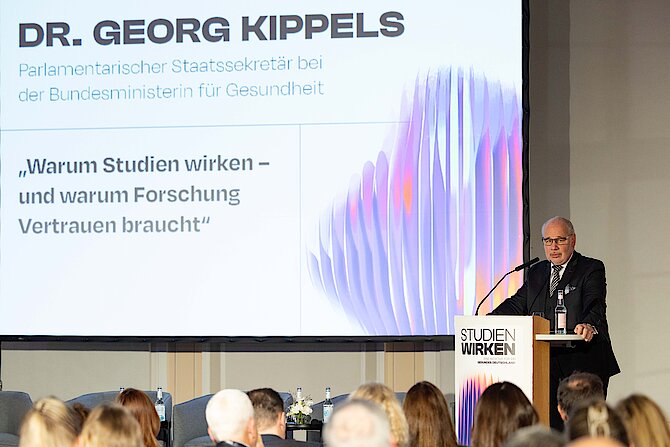 Mann steht am Rednerpult mit Mikrofon und Wasserflasche, im Hintergrund Projektion seines Vortrags, hier Vortragstitel und der Name des Vortragenden: Dr. Georg Kippels