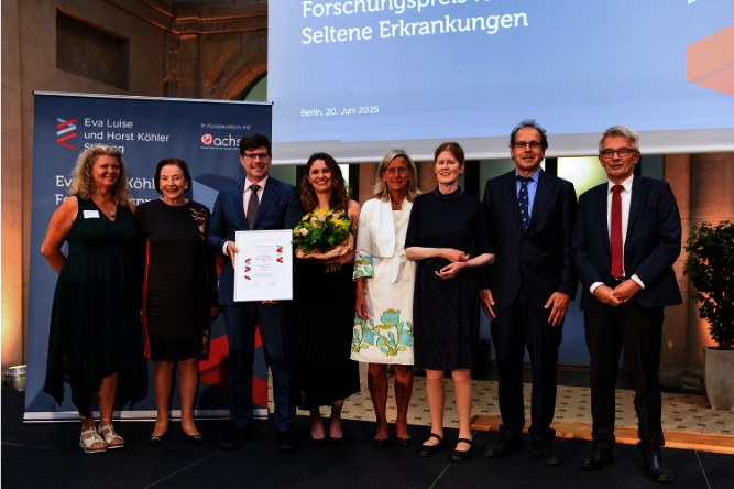 Gruppenbild der Gewinner des 16. Eva Luise Köhler Forschungspreises, Foto: Andrea Katheder Sieben Personen in festlicher Kleidung stehen nebeneinander auf einer Bühne, eine Person hält eine Urkunde, eine weitere einen Blumenstrauß, im Hintergrund ein Banner und eine Projektionsfläche mit Text.