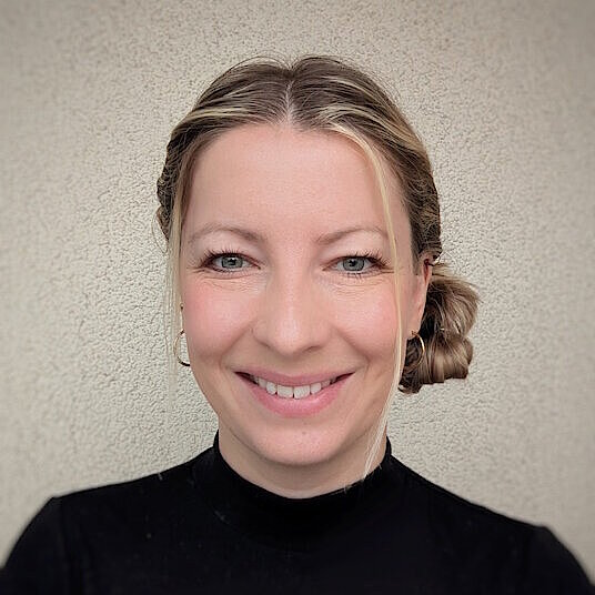 Person mit zusammengebundenem, hellbraunem Haar trägt schwarzen Pullover vor neutraler Wand.