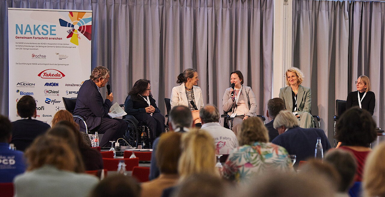 Fünf Personen sitzen auf einer Bühne vor einem Vorhang und diskutieren, davor sitzt ein Publikum.