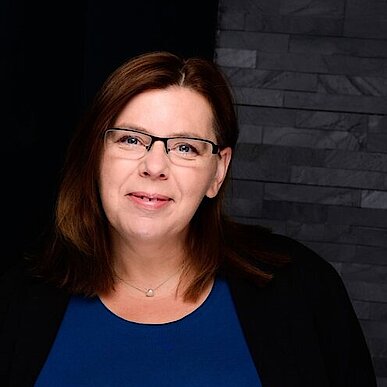 Nicole Heider Porträt einer Frau mit mittellangem braunen Haar, die eine Brille trägt. Sie hat ein blaues Oberteil und eine schwarze Jacke an und steht vor einem dunklen, strukturierten Hintergrund.