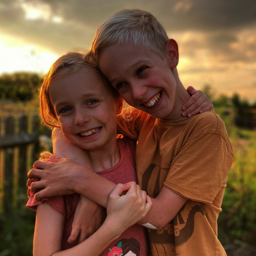 Ein Mädchen mit weggesteckten, langen blonden Haaren und ein Junge mit kurzen blonden Haaren umarmen sich draußen in einer ländlichen Umgebung bei Sonnenuntergang und lächeln in die Kamera. Bei genauerem Hinsehen ist erkennbar, dass der Junge eine Behinderung hat. Im Hintergrund sind ein Holzzaun und eine Wiese zu sehen, die in warmem Licht erstrahlen.