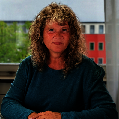 Geske Wehr Frau mit lockigem Haar und grünem Oberteil vor Fenster mit Blick auf grüne Bäume und rotes Gebäude