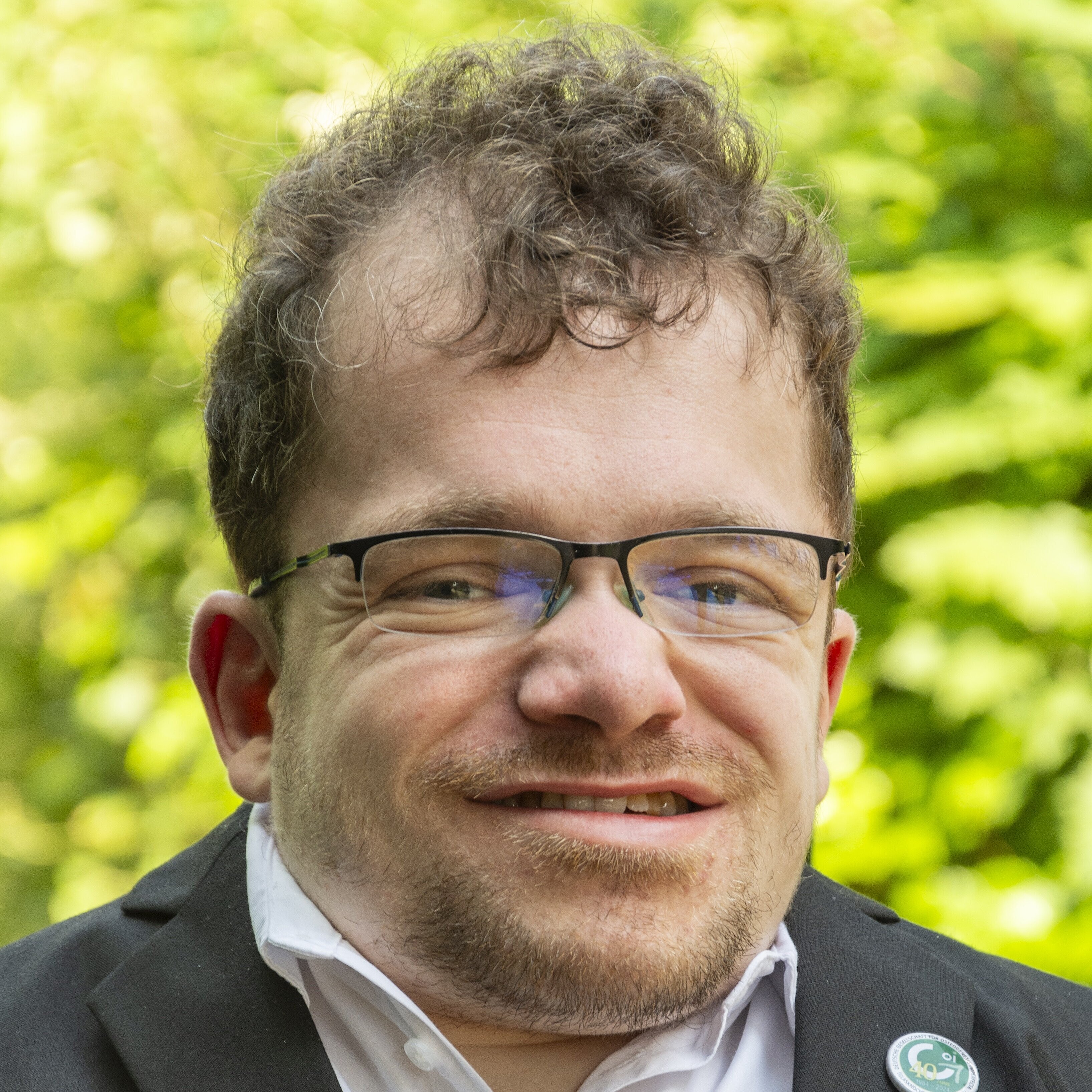 Porträt eines lächelnden Mannes im Freien mit lockigem braunem Haar, Brille und Bart. Er trägt einen dunklen Anzug mit weißem Hemd und einem runden Anstecker am Revers. Der Hintergrund ist unscharf und zeigt grünes Laub. Porträt eines lächelnden Mannes im Freien mit lockigem braunem Haar, Brille und Bart. Er trägt einen dunklen Anzug mit weißem Hemd und einem runden Anstecker am Revers. Der Hintergrund ist unscharf und zeigt grünes Laub.