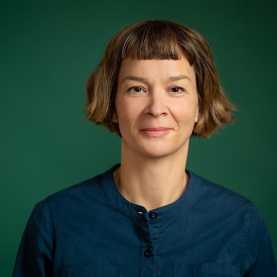 Person mit kurzen, gewellten braunen Haaren trägt ein dunkelblaues Hemd mit Stehkragen vor grünem Hintergrund.