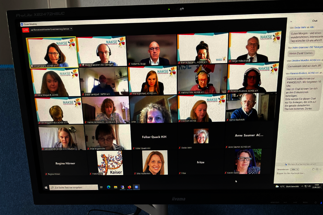 Computerbildschirm zeigt eine Videokonferenz mit mehreren Teilnehmern in Kacheln, darunter ein Chatfenster rechts, auf einem Schreibtisch mit Tastatur, Smartphone und Papieren.