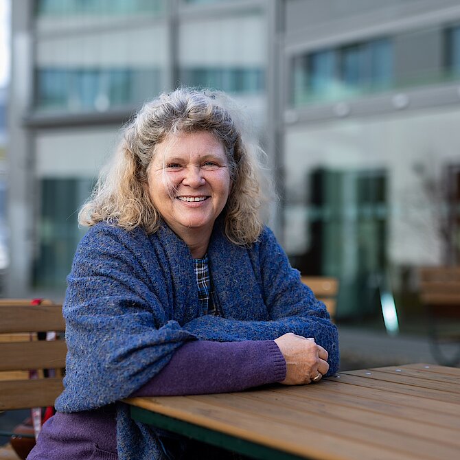 Person mit lockigem, schulterlangem Haar sitzt mit verschränkten Armen an einem Holztisch im Freien vor einem modernen Glasgebäude