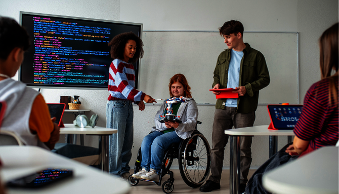 Drei junge Menschen, eine im Rollstuhl, zeigen in einem Klassenraum mit Computern und großem Bildschirm mit Programmiercode eine technische Konstruktion
