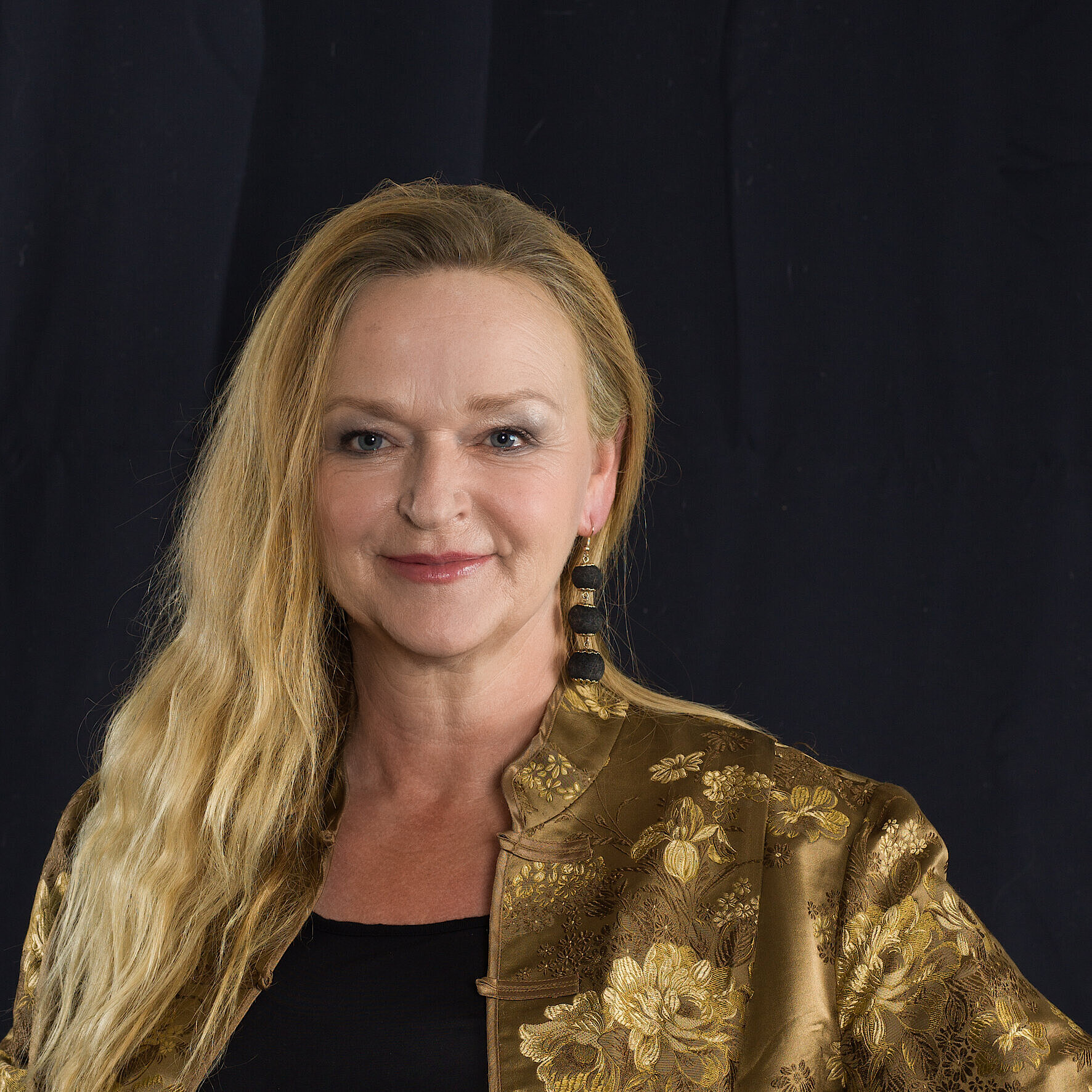 Porträt einer Frau mit langen, blonden Haaren, die leicht gewellt sind. Sie trägt eine goldfarbene, gemusterte Jacke und große schwarze Ohrringe vor einem dunklen Hintergrund.