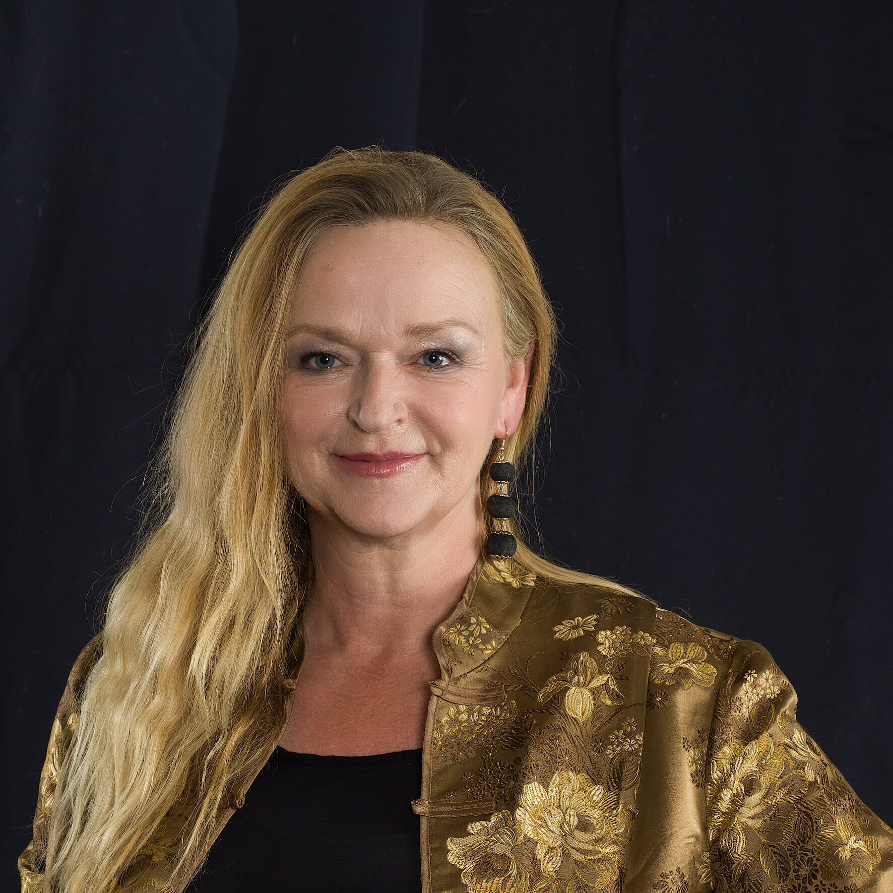 Porträt einer Frau mit langen, blonden Haaren, die leicht gewellt sind. Sie trägt eine goldfarbene, gemusterte Jacke und große schwarze Ohrringe vor einem dunklen Hintergrund.