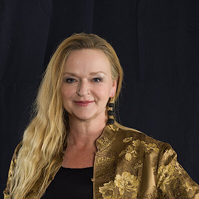 Porträt einer Frau mit langen, blonden Haaren, die leicht gewellt sind. Sie trägt eine goldfarbene, gemusterte Jacke und große schwarze Ohrringe vor einem dunklen Hintergrund.
