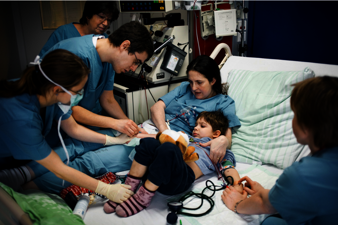 Mehrere Pflegekräfte betreuen ein Kind im Krankenhausbett, das ein Kuscheltier hält und an medizinische Geräte angeschlossen ist.