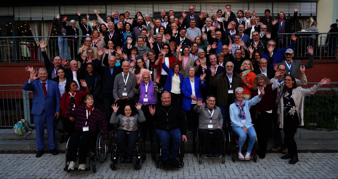 Große Gruppe von Menschen, darunter mehrere Personen im Rollstuhl, posiert vor einem mehrstöckigen Gebäude auf einer Treppe und winkt in die Kamera.