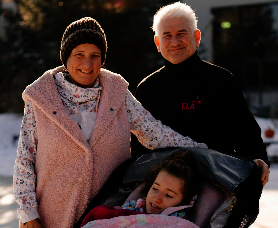 Drei Personen im Freien, eine Person sitzt in einem Kinderwagen mit einer Decke, die anderen beiden stehen daneben, eine trägt eine rosa Weste, die andere ein schwarzes Oberteil mit rotem Schriftzug