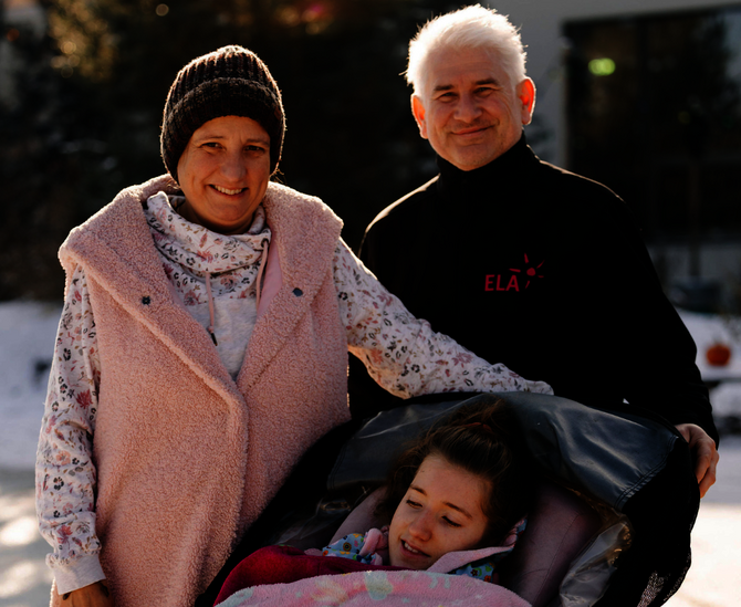Drei Personen im Freien, eine Person sitzt in einem Kinderwagen mit einer Decke, die anderen beiden stehen daneben, eine trägt eine rosa Weste, die andere ein schwarzes Oberteil mit rotem Schriftzug