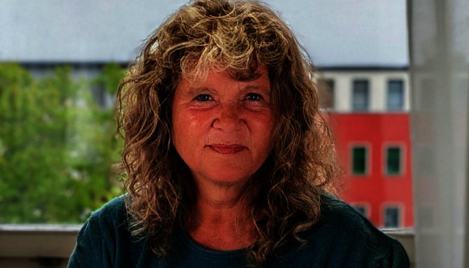 Frau mit lockigem Haar und grünem Oberteil vor Fenster mit Blick auf grüne Bäume und rotes Gebäude