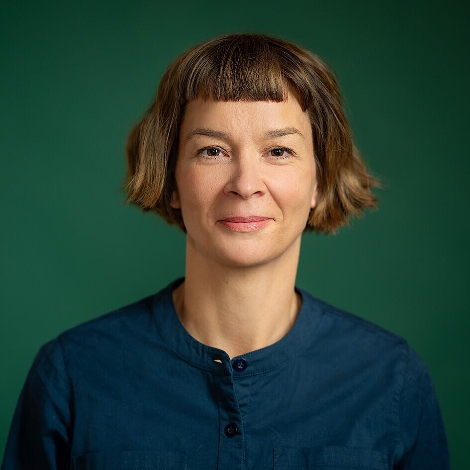 Person mit kurzen, braunen Haaren trägt ein dunkelblaues Hemd mit Stehkragen vor grünem Hintergrund.