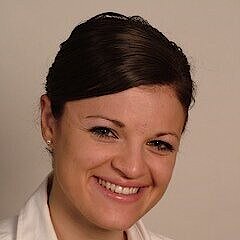 Frau mit dunklen, zurückgekämmten Haaren trägt weiße Bluse und kleine Ohrringe vor einfarbigem Hintergrund.