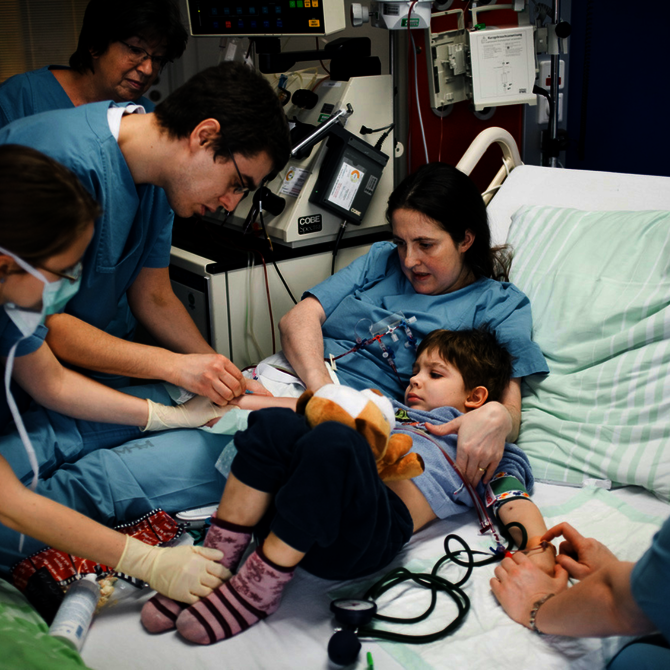 Mehrere Pflegekräfte betreuen ein Kind im Krankenhausbett, das ein Kuscheltier hält und an medizinische Geräte angeschlossen ist.