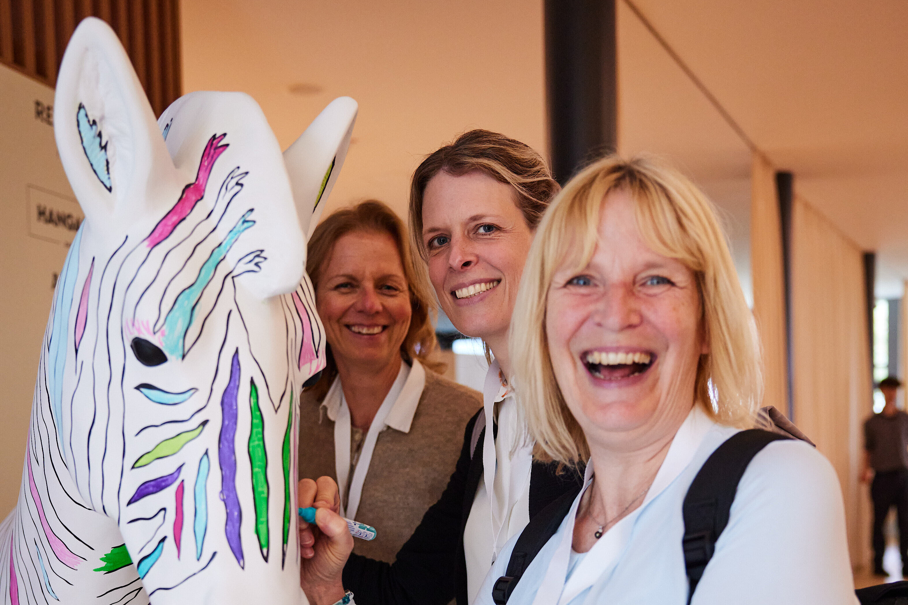 Lebensgroßes Zebra-Modell mit bunten, streifenförmigen Mustern auf weißem Grund steht auf grünem Podest vor Wand mit Wegweisern und Poster