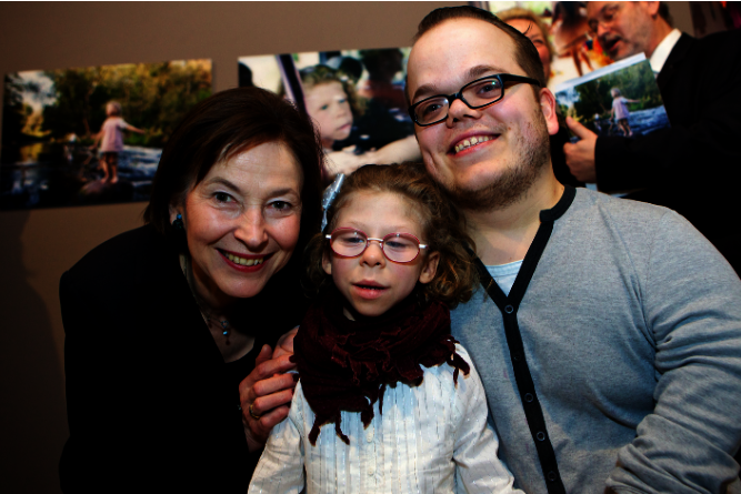 Drei Personen sitzen nah beieinander, eine trägt einen hellgrauen Pullover, eine andere einen schwarzen Anzug, im Hintergrund hängen Fotos an der Wand.