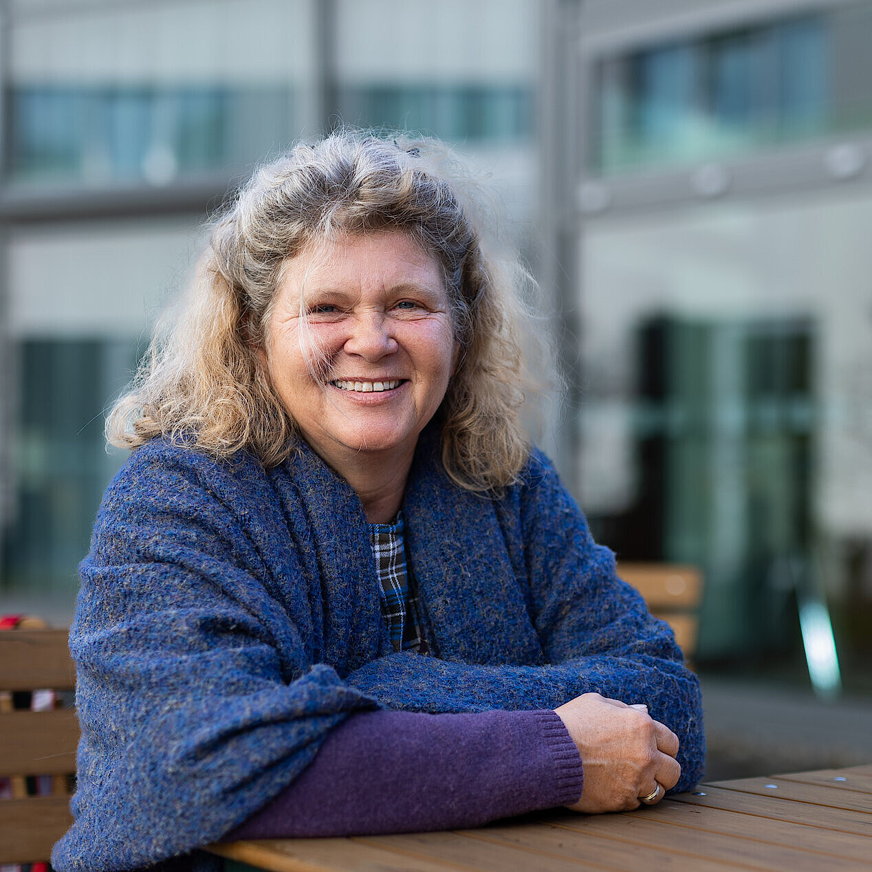 Person mit lockigem, schulterlangem Haar sitzt mit verschränkten Armen an einem Holztisch im Außenbereich vor einem modernen Gebäude.