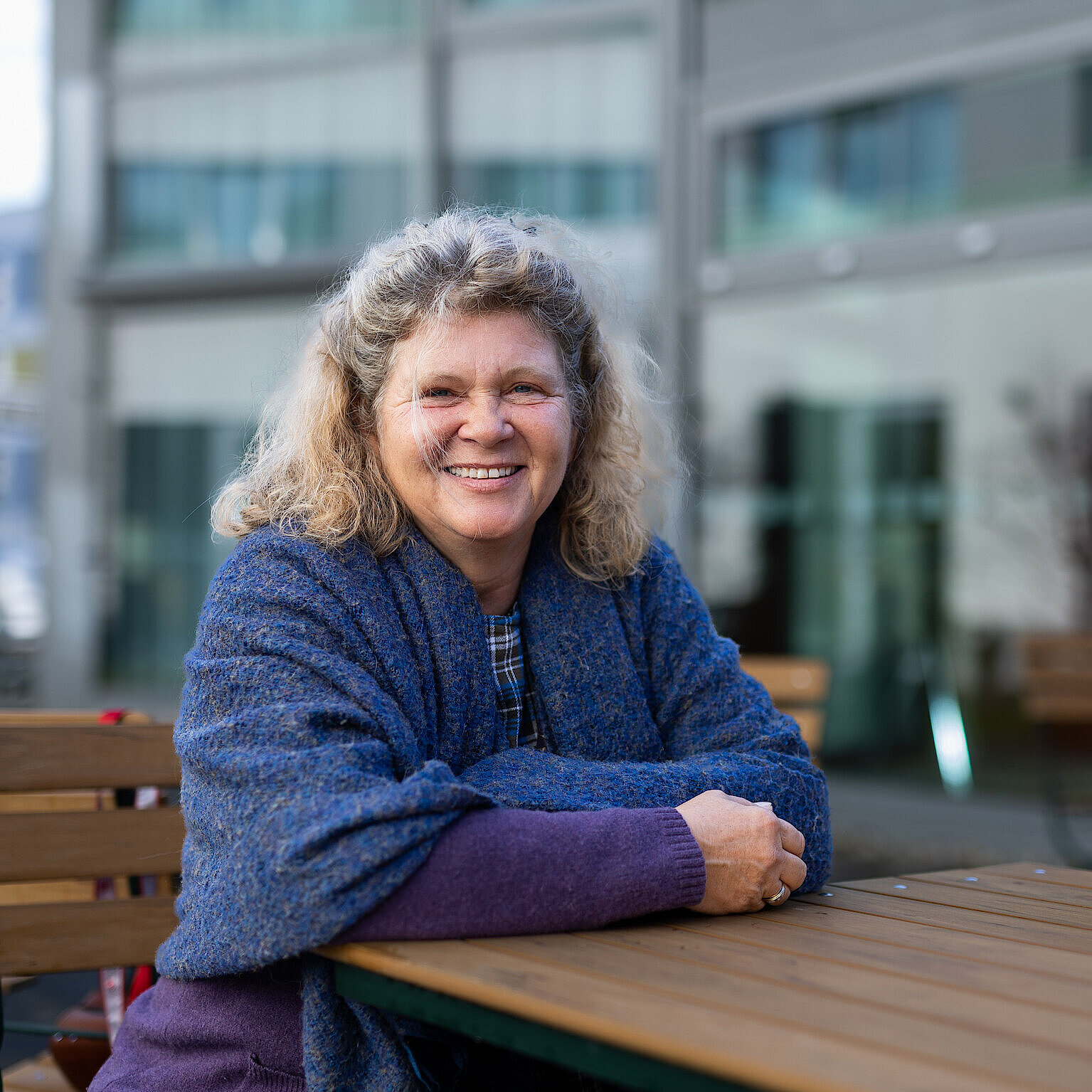 Person mit lockigem, schulterlangem Haar sitzt mit verschränkten Armen an einem Holztisch im Außenbereich vor einem modernen Gebäude.