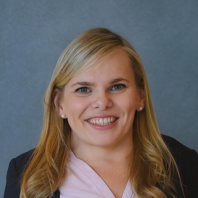 Frau mit langen blonden Haaren, trägt hellrosa Bluse und schwarzen Blazer vor blauem Hintergrund