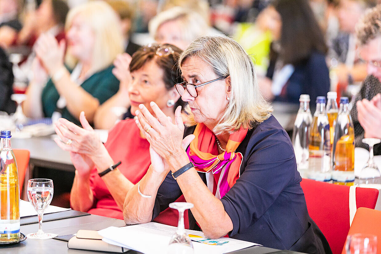 Mehrere Personen sitzen an Tischen und klatschen, im Vordergrund eine Frau mit einem bunten Schal und einem schwarzen Oberteil