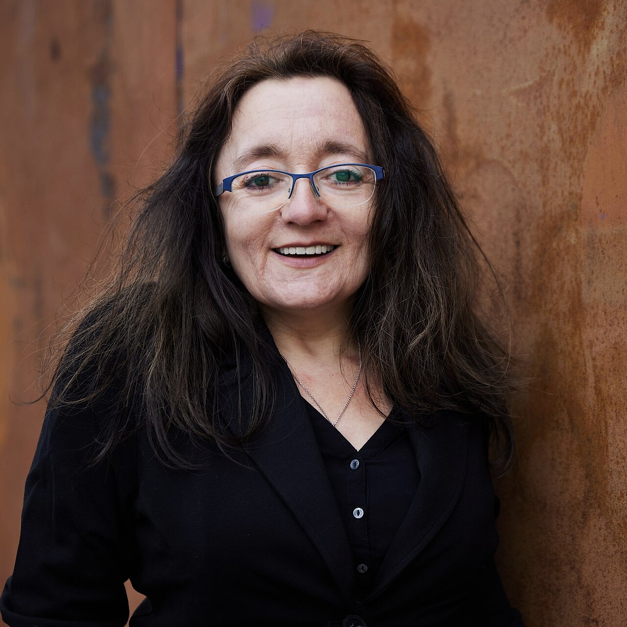 Frau mit langen, dunklen Haaren trägt schwarze Bluse und steht vor einer rostbraunen Wand