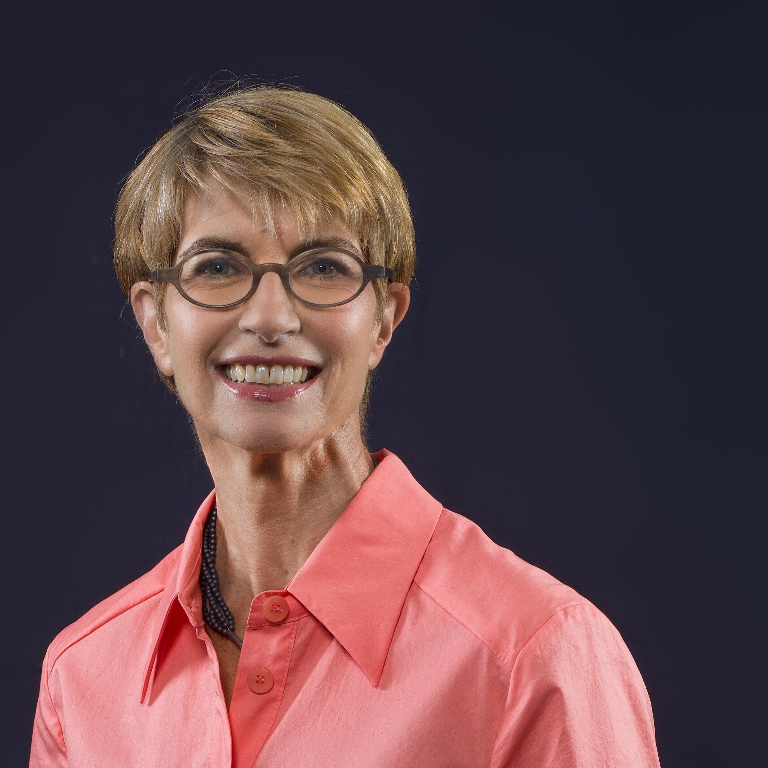 Person mit kurzem, blonden Haar trägt ein korallenrotes Hemd mit offenem Kragen und eine schwarze Halskette vor dunklem Hintergrund.