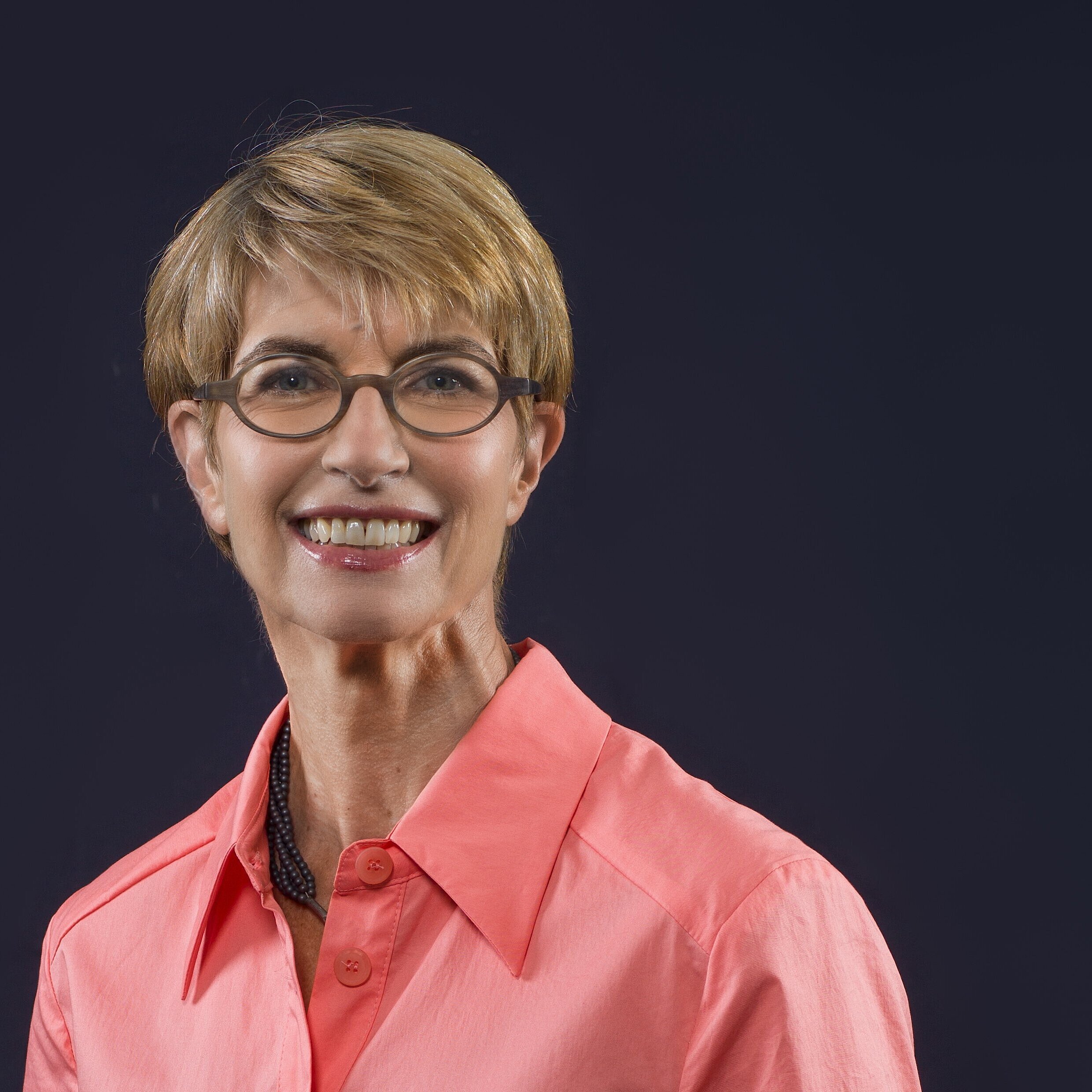 Person mit kurzem, blonden Haar trägt ein korallenrotes Hemd mit offenem Kragen und eine schwarze Halskette vor dunklem Hintergrund.