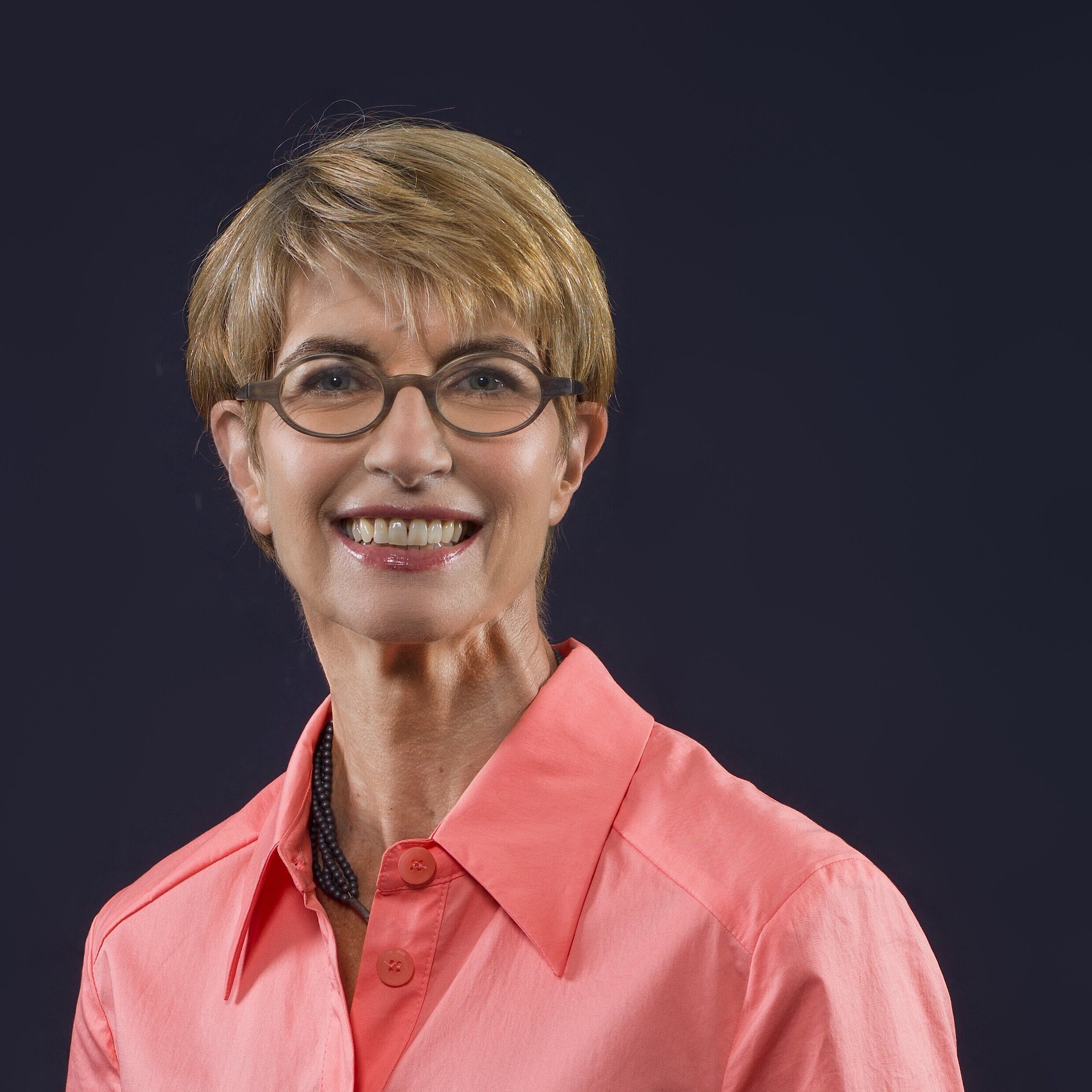 Person mit kurzem, blonden Haar trägt ein korallenrotes Hemd mit offenem Kragen und eine schwarze Halskette vor dunklem Hintergrund.