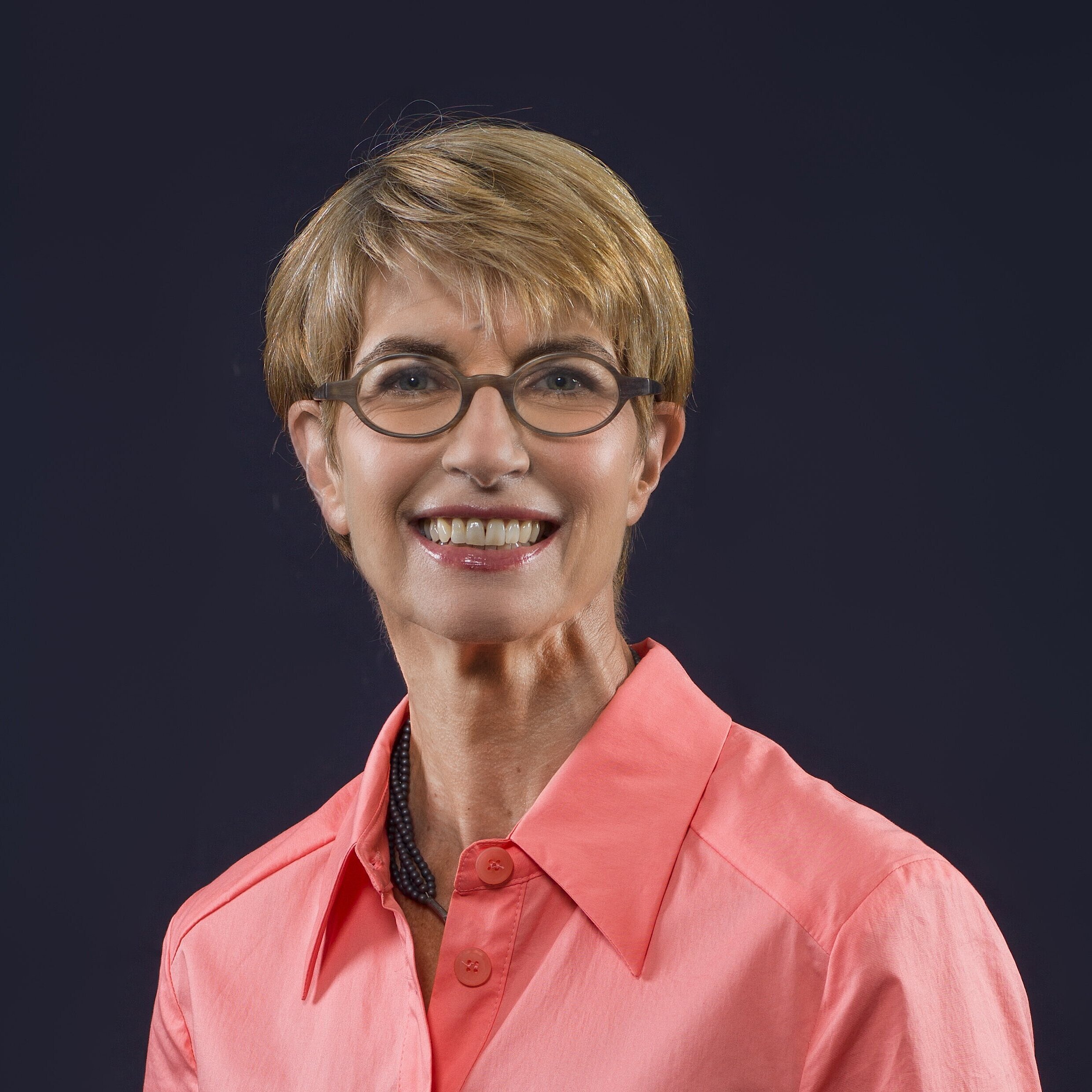 Person mit kurzem, blonden Haar trägt ein korallenrotes Hemd mit offenem Kragen und eine schwarze Halskette vor dunklem Hintergrund.