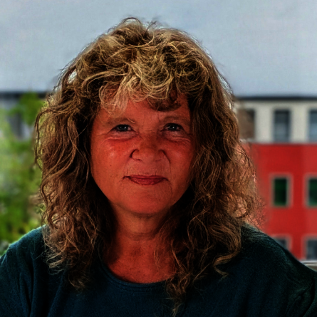Frau mit lockigem Haar, hellblauen Pullover über weißem Hemd vor einer hell verputzten Wand