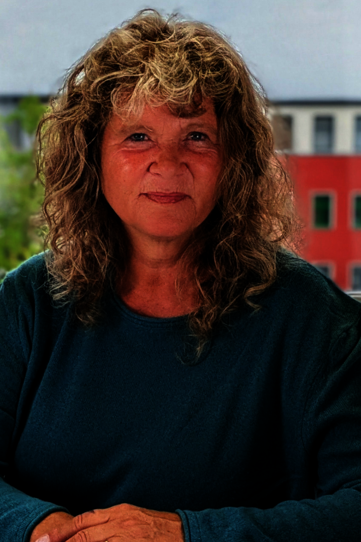 Frau mit lockigem Haar, hellblauen Pullover über weißem Hemd vor einer hell verputzten Wand